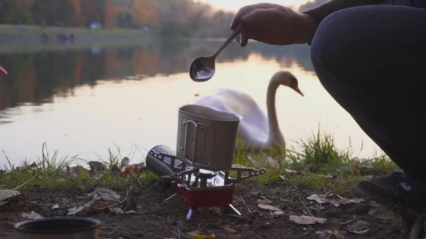 Camping food making. Tourist cooks coffee or tea or food on the gas stove. Beautiful landscape on the background of swans. Camp cooking on the shore of the lake and try the food. — Stock Video