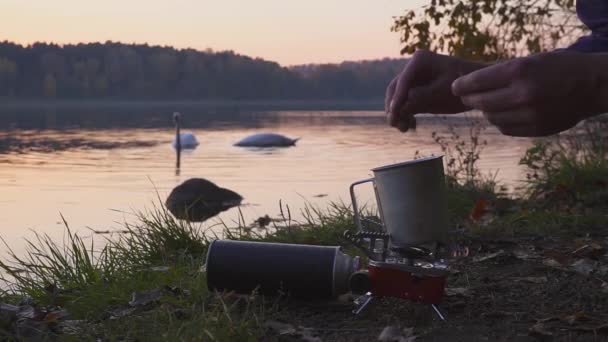 Camping food making. Tourist cooks coffee or tea or food on the gas stove. Beautiful landscape on the background of swans. Camp cooking on the shore of the lake. — Stock Video