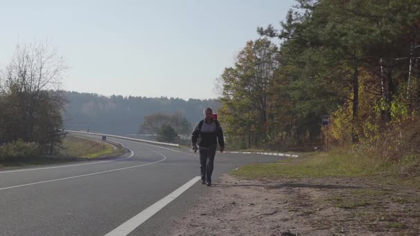 O homem vai numa estrada de asfalto com uma mochila nas costas. Outono Vista — Vídeo de Stock