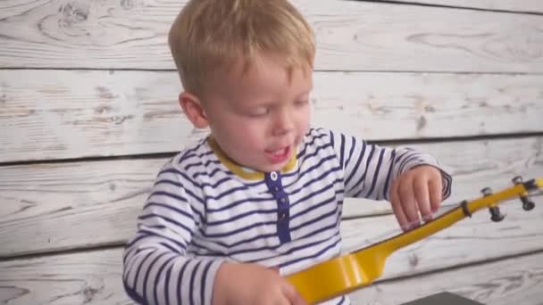 Feliz um ano de idade menino toca sua guitarra ou ukulele e canta canções, sentado na sala de madeira. — Vídeo de Stock
