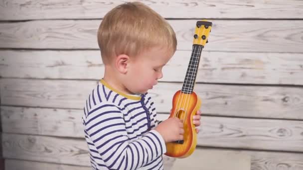 Feliz um ano de idade menino toca sua guitarra ou ukulele e canta canções, sentado na sala de madeira. — Vídeo de Stock