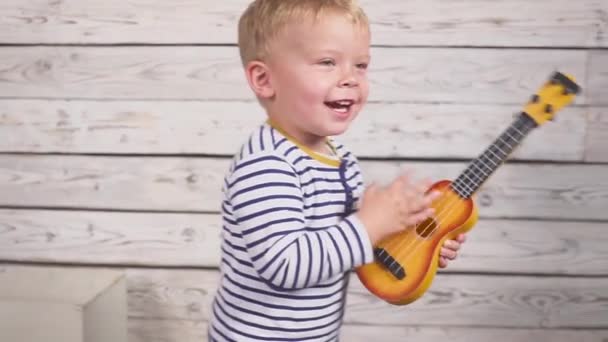 Glad en år gammal pojke spelar sin gitarr eller ukulele och sjunger låtar, sitter i trärummet. — Stockvideo