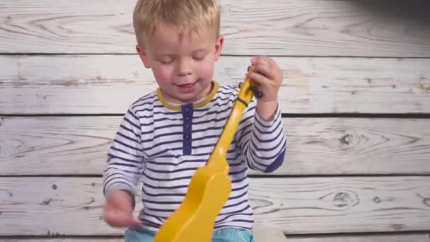 Feliz niño de un año toca su guitarra o ukelele y canta canciones, sentado en la habitación de madera. — Vídeos de Stock