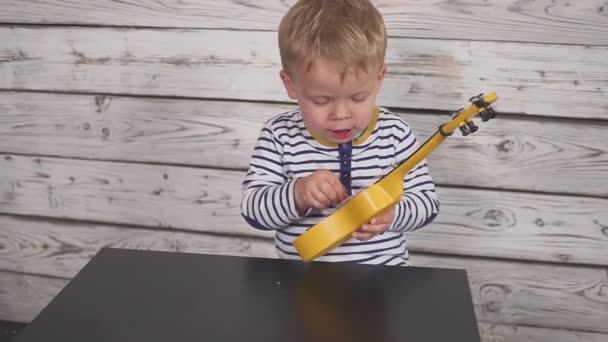 Щасливий однорічний хлопчик грає на гітарі або укулеле і співає пісні, сидячи в дерев'яній кімнаті . — стокове відео