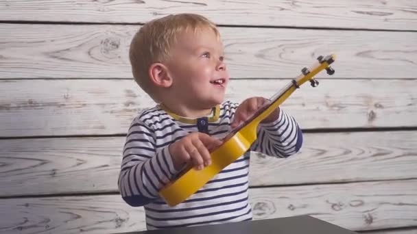 Feliz niño de un año toca su guitarra o ukelele y canta canciones, sentado en la habitación de madera. — Vídeo de stock