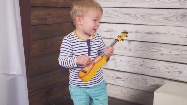 Boldog egy éves fiú gitározik vagy ukulele és dalokat énekel, ül a fa szobában. — Stock videók