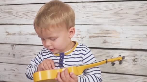 Один год мальчик играет на гитаре или укулеле и поет песни, сидя в деревянной комнате . — стоковое видео