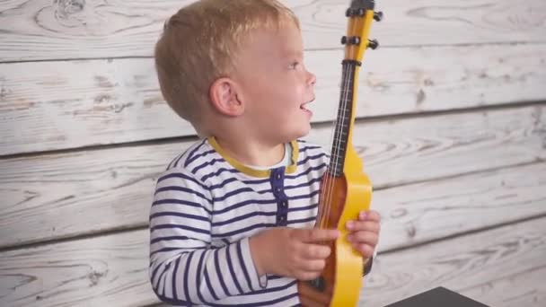 Un niño de un año toca su guitarra o ukelele y canta canciones, sentado en la habitación de madera . — Vídeo de stock