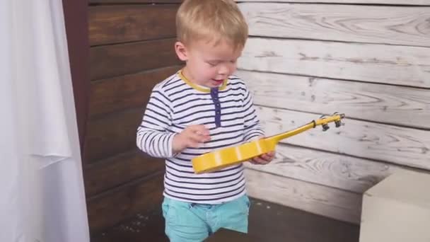 Un niño de un año toca su guitarra o ukelele y canta canciones, sentado en la habitación de madera . — Vídeo de stock