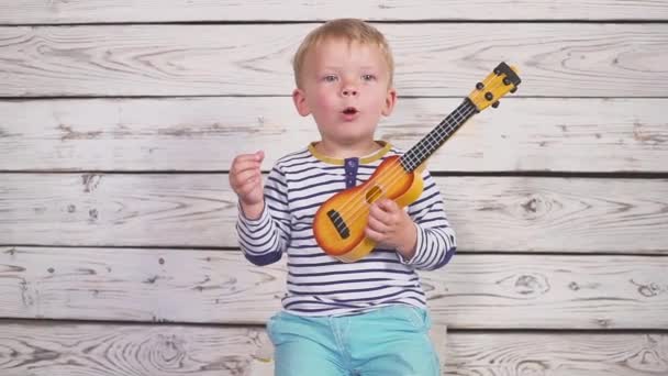 Un ragazzo di un anno suona la chitarra o ukulele e canta canzoni, seduto nella stanza di legno . — Video Stock