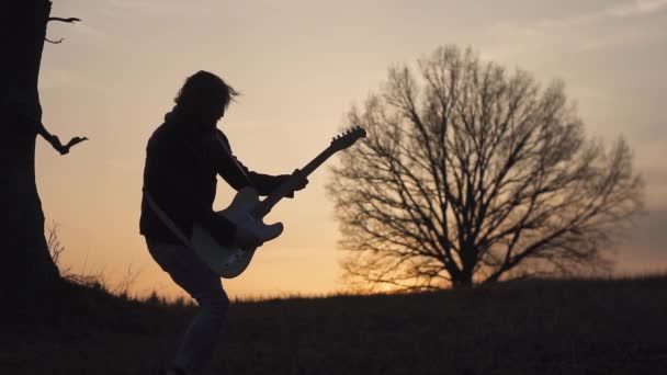 Az ember elektromos gitározni és énekelni az olyan területen, közel a fa a naplemente. sziluettjét — Stock videók