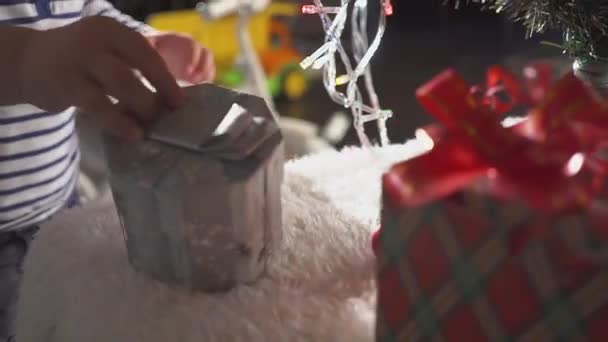 Niño dulce de dos años recibiendo un regalo con una cinta roja detrás de un árbol de Navidad con otras muchas cajas de regalo. Árbol de Navidad decorado con luces y bolas doradas. Vacaciones . — Vídeos de Stock