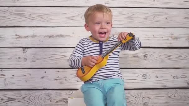 Niño feliz de dos años toca su guitarra o ukelele y canta canciones, sentado en la habitación de madera. — Vídeos de Stock