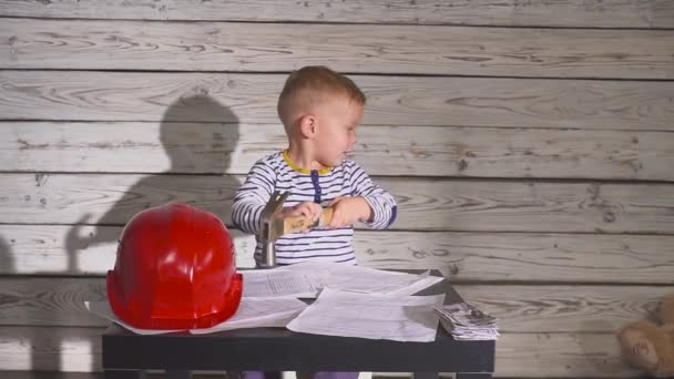 Cute Boy Constructor en ropa de trabajo y Helme. niño ingeniero trabajando en un proyecto con un martillo en sus manos. Sobre la mesa hay un casco, dinero en efectivo . — Vídeos de Stock