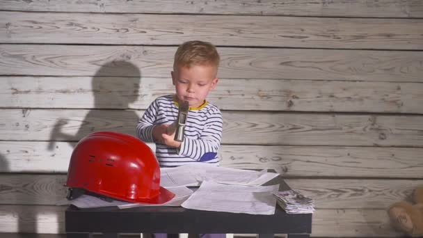 Leuke jongen Constructor In werkkleding en Helme. jongen ingenieur werken aan een project met een hamer in zijn handen. Op de tafel is een helm, contant geld. — Stockvideo