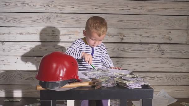 Χαριτωμένο αγόρι κατασκευή σε ρούχα και Helme εργασίας. αγόρι μηχανικός που εργάζονται για ένα σχέδιο με ένα σφυρί στα χέρια του. Στο τραπέζι είναι ένα κράνος, μετρητά χρήματα. — Αρχείο Βίντεο