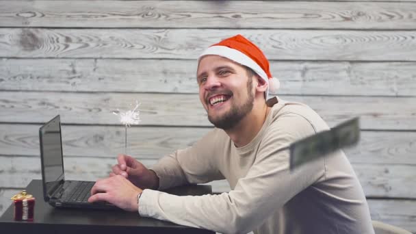 Dromerige man in een kerstmuts met Bengalen brand sparkler en laptop in zijn handen. Dalende dollar van het geld. — Stockvideo