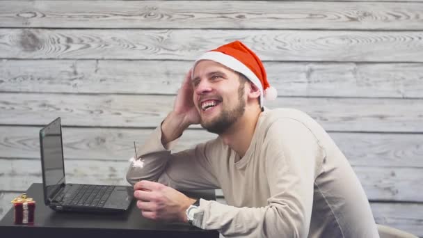Hombre guapo con símbolo de Santa es el uso de un ordenador portátil y se regocija con el fuego chispeante — Vídeo de stock