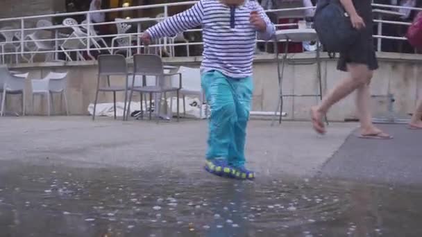 De jongen speelt in een plas in de regen. Hij verheugt zich in de regen. Twee-jaar-oude jongen uitgevoerd op de stoep. Concept op het thema van de droogte en warmte. stad — Stockvideo