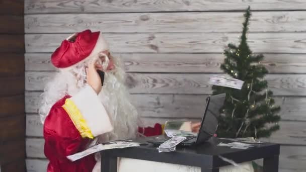 Санта Клаус говорит по мобильному телефону, работает за компьютером, и деньги падают на него — стоковое видео