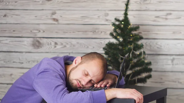 Ofiste genç işadamı yeni yıl arifesinde yorgun. Ofiste Noel ağacı. Laptop üstünde okul sırası. İş kavramı. Siyah elbiseli adam. Yeni yıl kutlama. Dijital aygıt kullanarak. Genç adam.