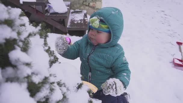 Niño el niño se encuentra en la nieve cerca del árbol de Navidad y se juega con decoraciones al aire libre . — Vídeos de Stock