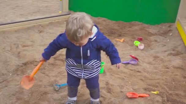 Jogos de crianças. Rapaz a cavar na caixa de areia. Sorri e toca a areia . — Vídeo de Stock