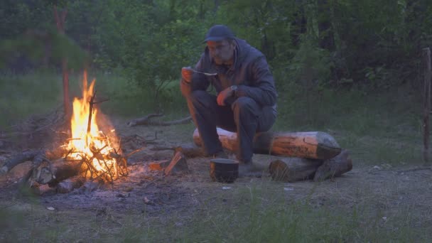 Mężczyzna podróżnik zjada z puli w pobliżu ognia w godzinach wieczornych — Wideo stockowe