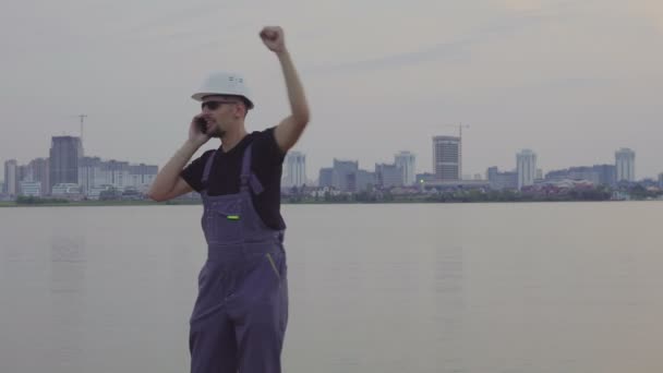 4 k의 아름 다운 저녁 도시 및 건설 현장 근처 휴대 전화에 흰색 헬멧에 잘생긴 백인 노동자 총. 건설 성공 — 비디오