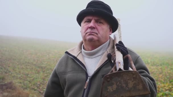 Granjero mayor con sombrero sosteniendo una azada fangosa y horquilla en el campo de primavera. Trabajo manual. Niebla — Vídeo de stock