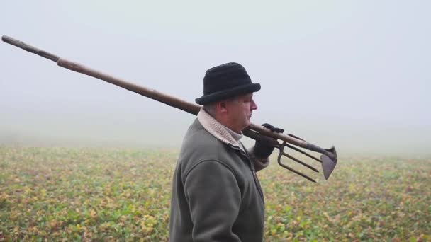 高级农民拿着帽子在春天的田野上拿着一个泥泞的锄头和叉子。体力劳动。雾 — 图库视频影像