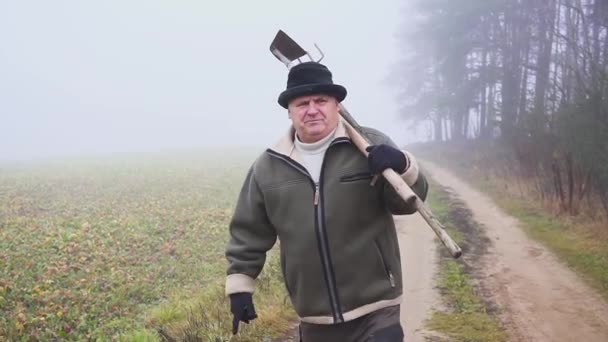 Senior bonde med hatt hålla en lerig hoe och högaffel på fältet. Kroppsarbete. Dimma — Stockvideo