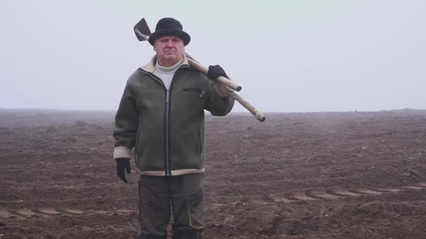 Ανώτερος γεωργός με καπέλο κρατώντας ένα λασπωμένο σκαπάνη και δίκρανο στο πεδίο. Χειρωνακτική εργασία. Ομίχλη — Αρχείο Βίντεο