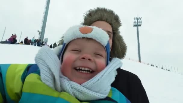 Ένας νεαρός άνδρας με ένα χαρούμενο αγόρι με ένα roller coaster βόλτα το χειμώνα στον πάγο. Όμορφος ο μπαμπάς και γιος γέλιο σε ένα πάρκο του χειμώνα. Ευτυχισμένη οικογένεια παίζει με το χιόνι. — Αρχείο Βίντεο