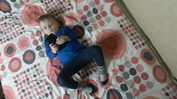 Niño acostado en la cama y mirando una pantalla de teléfono inteligente electrónico — Vídeos de Stock