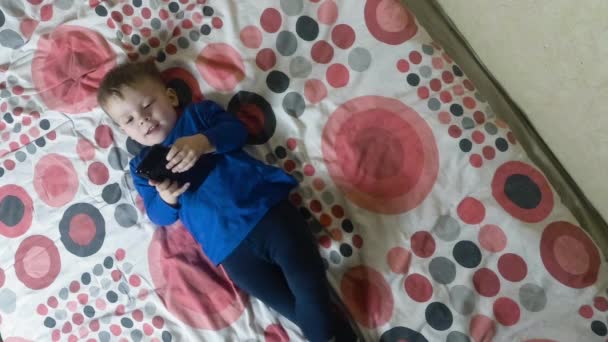 Niño acostado en la cama y mirando una pantalla de teléfono inteligente electrónico — Vídeos de Stock