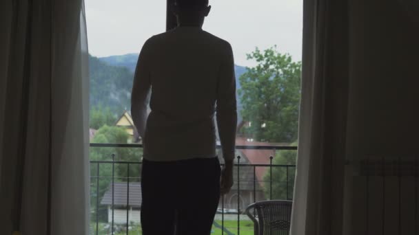 Νέος άνδρας πηγαίνει από το δωμάτιό του στο μπαλκόνι, Κοίτα στα βουνά. — Αρχείο Βίντεο