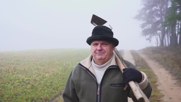 Senior bonde med hatt hålla en lerig hoe och högaffel på fältet. Kroppsarbete. Dimma — Stockvideo