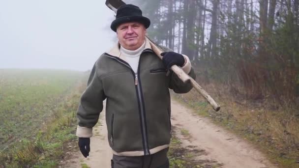 Старший фермер в шляпе, держащий грязную мотыгу и вилы на поле. Ручной труд. Туман — стоковое видео