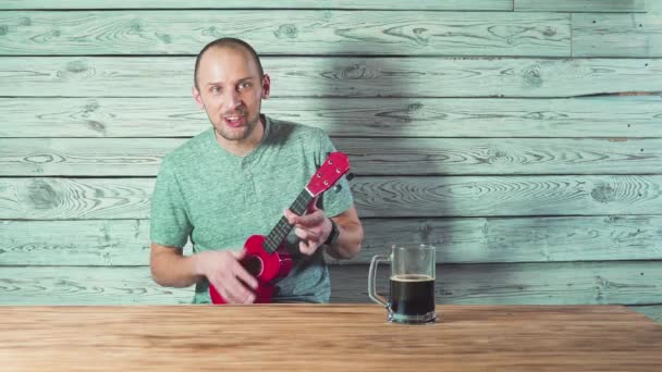 Młody człowiek pije piwo, śpiewając i grając ukulele dla swoich przyjaciół. — Wideo stockowe