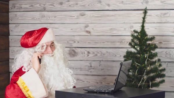 Papai Noel trabalhando com laptop em um escritório. Papai Noel usando um smartphone — Fotografia de Stock