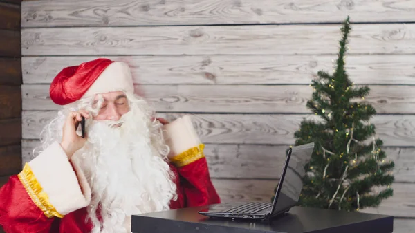 Papai Noel trabalhando com laptop em um escritório. Papai Noel usando um smartphone — Fotografia de Stock