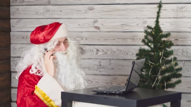 Ο Αϊ Βασίλης δουλεύει με λάπτοπ σε ένα γραφείο. Άγιος Βασίλης χρήση smartphone — Αρχείο Βίντεο