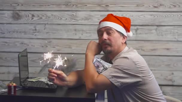 Verträumter Mann mit Weihnachtsmütze, Bengalfeuer-Wunderkerze und Laptop in der Hand. fallende Dollars. — Stockvideo