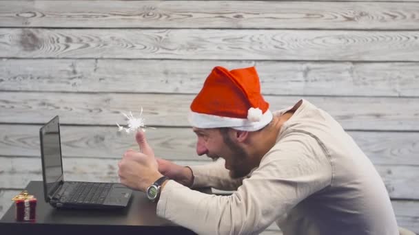 Hombre guapo con símbolo de Santa es el uso de un ordenador portátil y se regocija con el fuego chispeante — Vídeo de stock