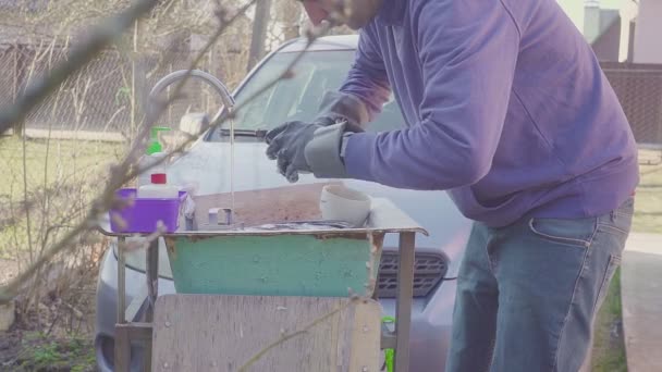 Joven lavando platos cerca del fregadero afuera. Las manos están frías de agua fría. El hombre con guantes. Vacaciones de camping. Concepto — Vídeo de stock