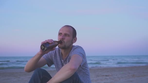 Um bêbado a beber cerveja na praia à noite. Retrato de um homem bebendo cerveja de uma garrafa — Vídeo de Stock