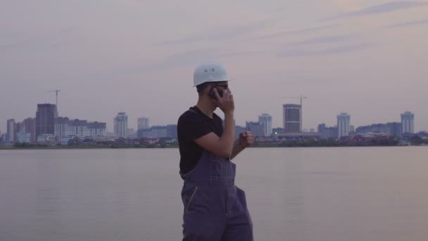 4K tiro de trabalhador caucasiano bonito em capacete branco falando no telefone celular perto da bela cidade da noite e canteiro de obras. sucesso na construção — Vídeo de Stock