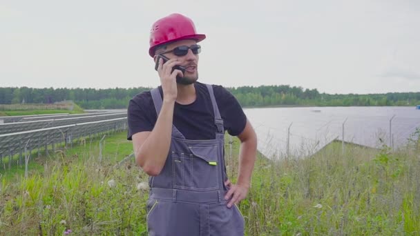 Retrato del trabajador que habla por teléfono cerca de una central solar y gestos, hace señales positivas — Vídeos de Stock