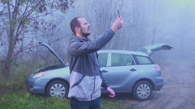adam kırık bir arabanın önünde bir mobil ağ arıyor. Sonbahar veya ilkbahar ormanı.